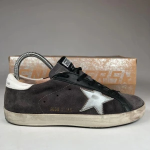 Grå mockasneakers med vit stjärna från Golden Goose - Säljer ett par grå sneakers från Golden Goose med vit stjärndetalj på sidan och svart snörning. Skorna har vit hälkappa och platt sula. Klassisk och avslappnad design med en cool vintagekänsla.