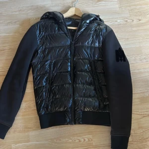  Mackage cardigan  - Snygg svart pufferjacka från Mackage med glansig framsida och huva. Jackan har dragkedja, två fickor fram och svarta ärmar i mjukare material. Perfekt för kyliga dagar och stilren look.