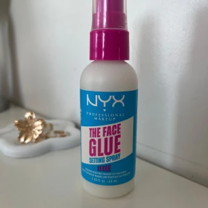 NYX The Face Glue Setting Spray - NYX Professional Makeup The Face Glue Setting Spray i en vit plastflaska med blå och rosa etikett. Sprayen har ett rosa lock och rymmer 60 ml. Perfekt för att fixera sminket hela dagen. Helt ny och oanvänd 