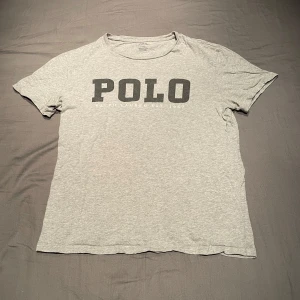 Grå t-shirt från Polo Ralph Lauren - Säljer en grå t-shirt från Polo Ralph Lauren med stor svart POLO-logga på bröstet. Klassisk rund halsringning och korta ärmar. Perfekt för en avslappnad stil. Pris kan diskuteras