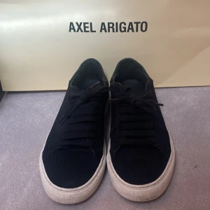 Svarta mocka sneakers från Axel Arigato - Snygga svarta sneakers från Axel Arigato med vit platt sula och diskret logga på sidan. Skorna har snörning och är tillverkade i skinn med en stilren och minimalistisk design. Perfekta för dig som gillar klassiska och trendiga sneakers. 