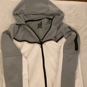 Nike teach  - Snygg hoodie från Nike i ljusgrått och vitt med svart dragkedja och logga på bröstet. Jackan har huva och lång ärm, samt en sportig design med svarta detaljer längs bröstet och ärmen.