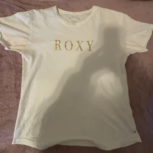 Vit t-shirt från Roxy - Säljer en vit t-shirt från Roxy i storlek XS. T-shirten har klassisk passform, rund halsringning och guldig ROXY-logga framtill. Perfekt basic-plagg med korta ärmar och mjukt bomullsmaterial. Finns svagt täcken på märke runt armhålan men skulle inte säga att det syns alls💗💕