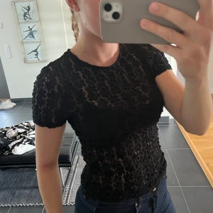 Svart spetsblus med kort ärm - Säljer en svart, tight spetsblus med kort ärm. Blusen har ett blommigt spetsmönster och rund halsringning. Perfekt att styla med jeans eller kjol för en trendig look.