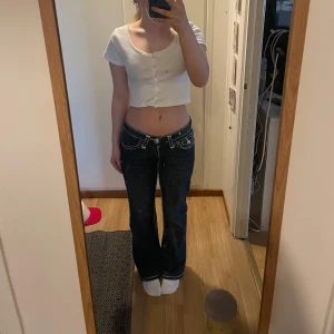 Mörkblå lågmidjade jeans från True Religion - Säljer ett par mörkblå jeans från True Religion med vita kontrastsömmar och stora fickor bak med dekorativa knappar. Jeansen har bootcut-modell och låg midja, perfekt för en avslappnad och trendig look. Storlek 28 33 som ni ser på sista bilden. Skriv om du har frågor eller vill ha fler bilder.