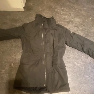 Svart parkajacka från Five Seasons - Svart parkajacka från Five Seasons med huva och dragkedja framtill. Jackan har flera fickor och en diskret logotyp på ärmen. Perfekt för kyligare dagar och designad i Sverige. Jacka som har aldrig använts ny pris 1100kr