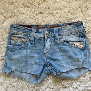 Ljusblå lågmidjade jeansshorts från Rock Revival - Säljer ett par ljusblå jeansshorts från Rock Revival med slitna detaljer, fransig kant och dekorativa bakfickor. Shortsen har silverfärgade knappar och dragkedja samt broderade detaljer på fickorna.