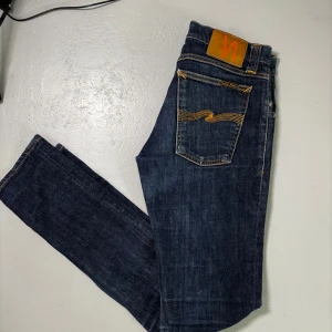  Nudie Jeans - Storlek.  :W28:L:32.                                        Skick:Topp skick.                              Beskrivning:Säljer ett par klassiska mörkblå jeans från Nudie Jeans med orangea kontrastsömmar och fem fickor. Jeansen har rak passform och normal midja, samt Nudie-logga på bakfickan och läderpatch bak i midjan.