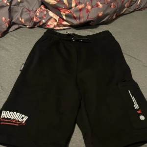Svarta shorts från Hoodrich med tryck - Svarta shorts från Hoodrich med vit och röd logga på benet och bakfickan. Resår i midjan med snörning och en ficka med dragkedja på sidan. Perfekta för en avslappnad stil.