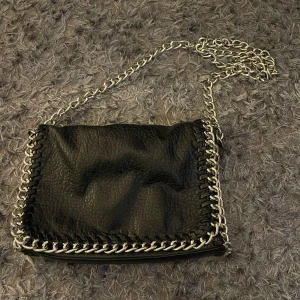 Svart axelväska med kedjedetaljer - Säljer en svart axelväska med grov silverfärgad kedja runt kanterna och som axelrem. Väskan har en cool design med flätade detaljer längs sidorna och är tillverkad i ett skinnliknande material.