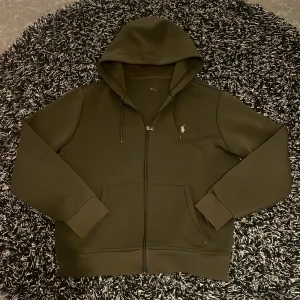 Olivgrön hoodie från Polo Ralph Lauren - Säljer en olivgrön hoodie från Polo Ralph Lauren med dragkedja och klassisk broderad logga på bröstet. Tröjan har långa ärmar, huva med dragsko och två fickor framtill. Perfekt för en avslappnad stil.