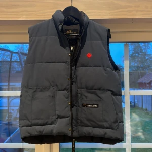 Mörkblå dunväst från Canada Goose - Säljer en mörkblå dunväst från Canada Goose med röd lönnlöv-detalj på bröstet. Västen har två stora fickor framtill, dragkedja och knappar. Perfekt för kyliga dagar när du vill hålla dig varm och snygg.