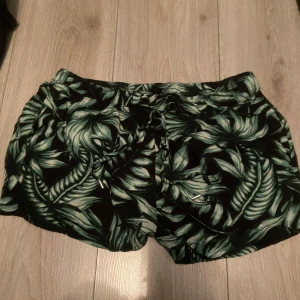 Svarta shorts med grönt blad-mönster från H&M - Snygga svarta shorts från H&M med tropiskt grönt blad-mönster. De har elastisk midja med snörning och är tillverkade i ett lätt och bekvämt material, perfekta för varma dagar. Passar s också 