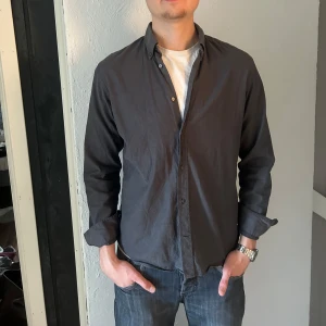 Mörkgrå skjorta från J Lindberg  - Säljer en stilren mörkgrå skjorta från J Lindberg. Skjortan har klassisk krage, knäppning framtill och långa ärmar. Perfekt till jeans eller chinos för en avslappnad men snygg look.