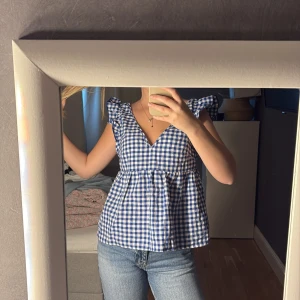 Blåvit rutig blus med öppen rygg - Supersöt blus i blåvitt rutigt mönster med volangdetaljer vid axlarna och djup v-ringning. Toppen har öppen rygg med knytband och är perfekt till sommaren. Luftig och lätt att matcha med jeans eller kjol.