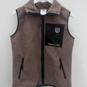 Brun fleeceväst från Maya Delorez - Mysig brun fleeceväst med svarta detaljer och dragkedjor. Västen har broderad logga på bröstet och tryck på ryggen med Jönköping Horse Show. Perfekt för ridning och stallhäng. 