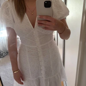 Vit broderad klänning med puffärm och knappar - Superfin vit klänning med broderade blommor, puffärmar och v-ringning. Knappar framtill och volang nertill ger en romantisk vibe. Perfekt för sommarens alla tillfällen!