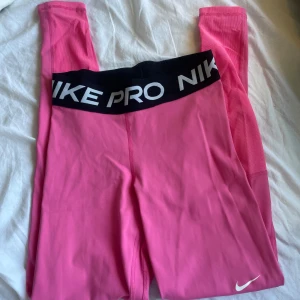 Rosa träningstights från Nike Pro - Säljer ett par rosa Nike Pro träningstights med svart resår i midjan där det står 'NIKE PRO'. Tightsen har en vit Nike-logga på benet och är tillverkade i Dri-FIT material som andas. Perfekta för gym eller löpning.