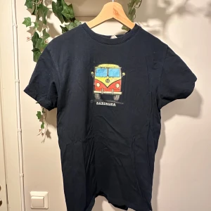 Mörkblå t-shirt med biltryck från Sardegna - Säljer en mörkblå t-shirt med ett färgglatt tryck av en klassisk bil och texten 'Sardegna' på bröstet. Den är köpt i Sardinien. T-shirten har rund hals och korta ärmar, perfekt för en avslappnad stil. Sitter som en S
