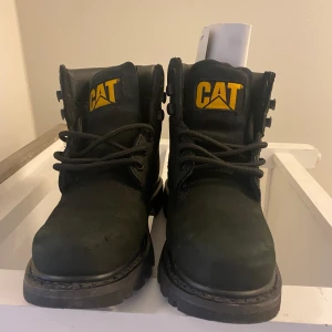 Svarta boots från Caterpillar - Tunga svarta boots från Caterpillar med högt skaft och kraftig sula. Skorna har snörning framtill och tydlig gul logotyp på plösen och sidan. Perfekta för dig som vill ha en robust och stilren look.