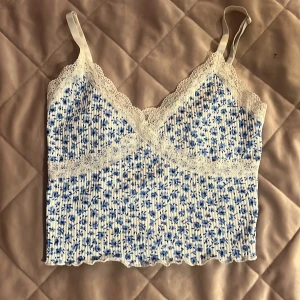 Blommig croppad linnetopp med spets från H&M Divided - Söt croppad linnetopp från H&M Divided med blått blommönster på vit botten. Toppen har smala axelband, spetsdetaljer längs urringning och under bysten samt ribbad struktur. Perfekt för varma dagar!