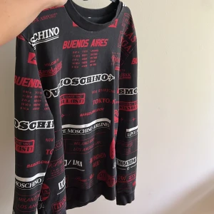 Svart sweatshirt från Love Moschino med röd och vit text - Cool svart sweatshirt från Love Moschino med tryck i rött och vitt över hela tröjan. Texten har inspiration från flygplatser och resor. Rund halsringning och långa ärmar. Perfekt statement-plagg för dig som gillar streetwear.