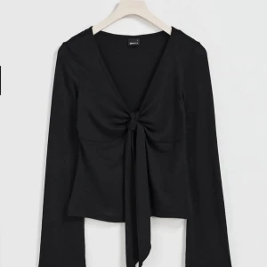 Svart långärmad topp med knyt framtill - Snygg svart långärmad topp med djup v-ringning och knyt framtill. Perfekt att styla med jeans eller kjol för en trendig look. Tunn och mjuk i materialet.