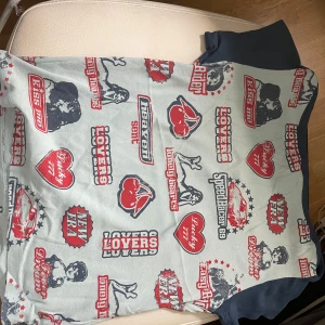 Mönstrad off shoulder t-shirt från Urban Outfitters - Snygg ljusblå off shoulder cropped t-shirt med  marin blåa ärmar och coola motiv. Tröjan e helt ny utan lapp. Jag skulle säga att tröjan passar storlek M men sitter även helt okej på storlek S.