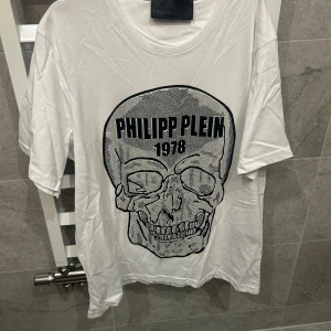 Vit t-shirt med döskalletryck från Philipp Plein - Säljer en vit t-shirt från Philipp Plein med stort svart döskalletryck och texten 'PHILIPP PLEIN 1978' på bröstet. T-shirten har rund halsringning och är kortärmad. Perfekt statement-plagg för dig som gillar unika designer.