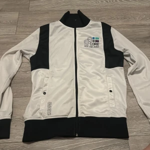 Vit och svart jacka från Jack & Jones - Säljer en sportig vit och svart jacka från Jack & Jones med dragkedja framtill och tryckt logga på bröstet. Jackan har långa ärmar, ståkrage och svarta detaljer vid ärmslut och midja. Perfekt för en avslappnad stil.