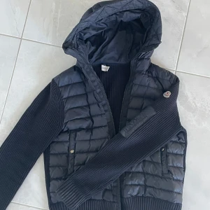 Svart pufferjacka med stickade ärmar från Moncler - Säljer en svart pufferjacka från Moncler med stickade ärmar och huva. Jackan har dragkedja framtill, två fickor med knappar och Moncler-logga på ärmen. Perfekt för kyliga dagar och riktigt snygg till vardags.