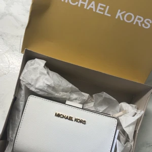 Vit plånbok från Michael Kors - Snygg och stilren vit plånbok från Michael Kors i läder. Plånboken har flera kortfack, ett fönster för ID, samt ett fack med dragkedja för mynt. Perfekt för att hålla ordning på dina kort och kontanter. Helt ny med allt som tillkom vid köp. Köptes för 1400kr. 