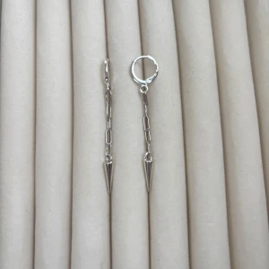 Silverfärgade örhängen med kedja och kon - Säljer ett par trendiga silverfärgade örhängen med tunn kedja och spetsig kon längst ner. Örhängena har en minimalistisk och edgy design som passar perfekt till dig som gillar stilrena accessoarer.