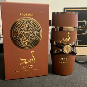 Asad Bourbon Eau de Parfum från Lattafa - Exklusiv parfym Asad Bourbon från Lattafa i en brun flaska med gulddetaljer och orientaliskt mönster. Flaskan har en unik form med metallisk dekor och matchande ask. Doften är inspirerad av bourbon och har lyxig känsla.
