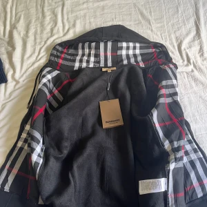 Svart hoodiejacka från Burberry - Säljer en svart hoodiejacka från Burberry med klassiskt rutigt foder i huvan och insidan. Jackan har dragkedja framtill, snörning i huvan och broderad logga på bröstet. Perfekt för dig som gillar stilrena och exklusiva plagg.