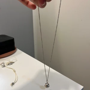 Halsband med fotbollsberlock - Snyggt halsband med en kedja i silverfärg och en berlock formad som en fotboll i vitt och svart. Perfekt accessoar för dig som älskar fotboll och vill visa det på ett stilrent sätt. Kedjan är tunn och smidig, och berlocken är rund och detaljerad.