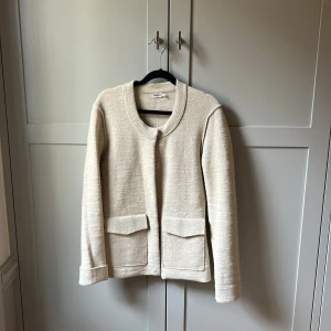Beige cardigan kavaj från Filippa K - Stilren beige cardigan jacka från Filippa K med rundad krage, två stora fickor framtill och diskreta knappar. Jackan är i ett mjukt, stickat material och har långa ärmar. Perfekt för dig som gillar minimalistisk och clean stil.