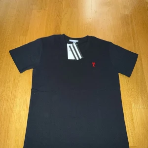 Svart t-shirt från Ami - Snygg svart t-shirt från Ami med ett rött broderat hjärta och bokstaven A på bröstet. Perfekt för en stilren look. Gjord i mjukt bomullsmaterial och har en klassisk passform. Priset kan diskuteras 