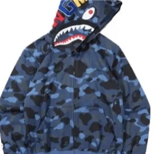 BAPE hoodie  - Säljer en fet BAPE Shark vindjacka med blått camo-mönster och ikonisk hajdesign på huvan. Jackan har dragkedja framtill och broderade bokstäver på huvan. Perfekt för dig som vill sticka ut med streetwear-vibe och coola detaljer.