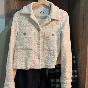 Vit  cardigan jacka från Zara - Säljer en vit  cardigan jacka från Zara med snygga guldfärgade knappar och två stora fickor framtill. Jackan har en cool textur i stickad polyester och klassisk krage. Perfekt för dig som gillar trendiga och stilrena plagg.