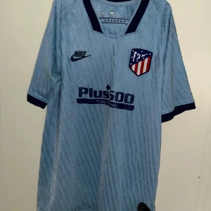 Blå Atlético Madrid Nike fotbollströja - Snygg ljusblå fotbollströja från Nike med Atlético Madrid-emblem på bröstet och Plus500-tryck. Tröjan har korta ärmar med mörkblå muddar, V-ringad krage och diskret randigt mönster. Tillverkad i lätt Dri-Fit material som andas och passar perfekt för träning. Tröjan har små fläckar som förmodligen går att tvätta bort.