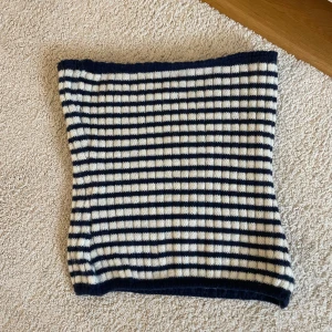 Randig stickad halsduk i blått och vitt - Snygg  tubtopp med randigt mönster i marinblått och vitt. Stickad i mjukt material som håller dig varm under kyliga dagar. Perfekt att slänga på sig för en cool och avslappnad look.