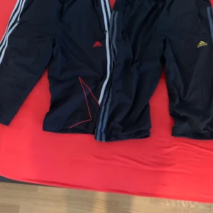 Svarta Adidas träningsshorts långa M - Två par svarta Adidas träningsshorts med klassiska ränder längs sidorna. Ena paret har röd logga och röda detaljer, det andra har gul logga. Båda är i lätt och ventilerande material, perfekta för sport eller chill. Elastisk midja och raka ben. 