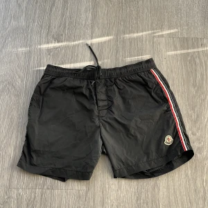 Svarta Moncler badshorts med snörning - Svarta badshorts från Moncler med klassisk logga på benet och snygga rödvita ränder längs sidorna. Shortsen har elastisk midja med snörning och meshfoder på insidan. Perfekta för sommaren och chill vid poolen.
