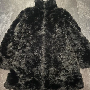 Svart pälsjacka från Softy, stl S - Fluffig svart pälsjacka från Softy med hög krage och dragkedja framtill. Jackan har en oversized känsla och är perfekt för kalla dagar när du vill vara både varm och snygg. Mjuk och gosig med lyxig look. Jackan har ett knytband som tyvärr är borta.