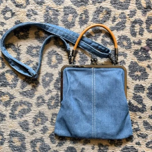 Snygg blå denimbäska med snäppspänne  - Jättesnygg blå denim handväska med ställbar axelrem och ett ljusbrunt läderhandtag Väskan stängs & öppnas med ett snäppspänne Den har ett fint blå/vitt randigt innerfoder med ett stort fack och ett litet fack med dragkedja  Som ny i jättefint skick! Mått: H21,5/B23 cm