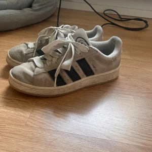 Adidas Campus sneakers i mocka - Snygga Adidas Campus sneakers i beige mocka med klassiska svarta ränder på sidorna och vit sula. Skorna har rund tå, platt sula och breda vita snören. Perfekta för dig som gillar retrostil och streetwear. (Inte rengjorda på bilderna) strl 40 men mer som 39