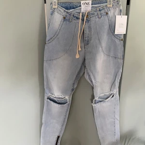 Ljusblå jeans med snörning från ONE teaspoon  - Snygga ljusblå jeans från ONE teaspoon med avslappnad passform, snörning i midjan och slitna hål på knäna. Jeansen har dragkedjedetalj vid benslut och bakfickor med unik form. Perfekta för en cool och avslappnad stil.