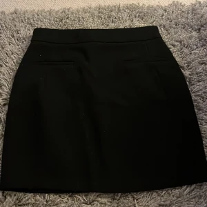 Svart kortkjol med fickdetaljer - En stilren svart kortkjol med två diskreta fickdetaljer framtill och dold dragkedja bak. Kjolen har en klassisk rak passform och är perfekt för dig som gillar en clean och enkel look. Materialet känns mjukt och följsamt.