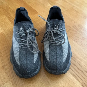 Grå  svarta sneakers från Philipp Plein - Snygga sneakers från Philipp Plein i grått och svart med stickad ovandel och coola logodetaljer både fram och bak. Skorna har snörning och en chunky sula som ger en sportig vibe. Perfekta för dig som gillar streetstyle och vill sticka ut.